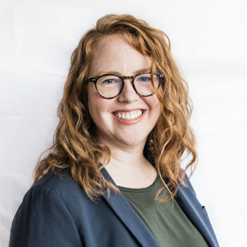Alexa Milton is a roughly 40-year-old white woman with wavy red hair and glasses. She is wearing a dark green shirt and navy suit jacket and is smiling.
