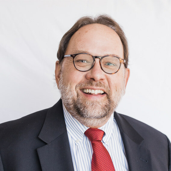 Michael Allen is a white man in his mid-60s with a beard and glasses. He is wearing a dark blue suit and a red tie and is smiling.