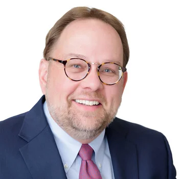 Michael Allen is a white man in his mid-60s with a beard and glasses. He is wearing a dark blue suit and a red tie and is smiling.