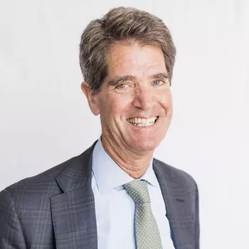 John Relman is a white man in his late sixties. He is wearing a blue suit and a green tie and is smiling.