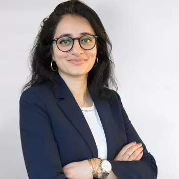 Ellora Israni is a South Asian woman in her 30s. She is wearing a blue jacket and glasses and smiling.