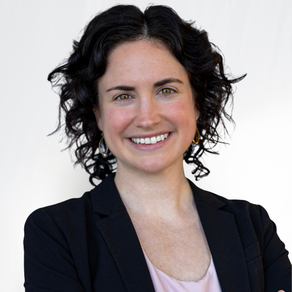 Brenna Kearns is a white woman in her 30s with brown hair. She is wearing a dark suit and metallic earrings and is smiling.